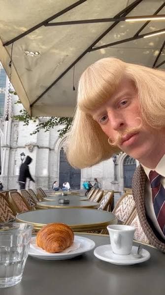 Vertical still of Jean Phil seated at a Paris cafe terrace in a brown houndstooth suit, blonde bob and curled mustache, with a croissant and espresso on the table